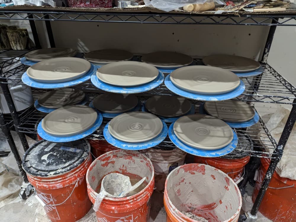 Abby's freshly thrown plates drying on studio shelves above the glaze buckets